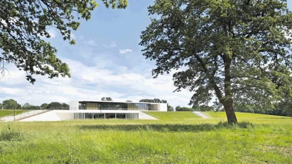 Krajinski pogled na &scaron;portni objekt NZS, kjer je  optično izpostavljen samo paviljon, ki percepcijsko lebdi nad zelenim nasipom.