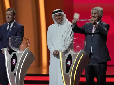 Former German soccer international and manager Lothar Matthaus, left, and former Qatari soccer international Adel Ahmed MalAllah look on as Serbian-Mexican retired soccer manager Bora Milutinovic holds up the name of Canada as he assists in the 2022 soccer World Cup draw at the Doha Exhibition and Convention Center in Doha, Qatar, Friday, April 1, 2022. (AP Photo/Darko Bandic)