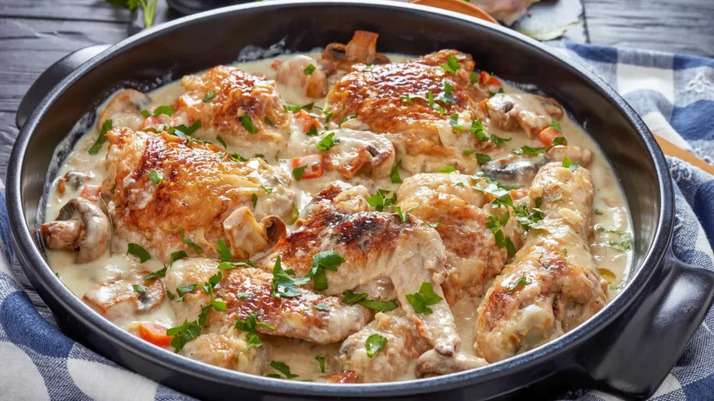 hot chicken fricassee in a black dutch oven on a black table - chicken meat browned and stewed in white wine cream sauce with mushrooms and vegetables, authentic french recipe