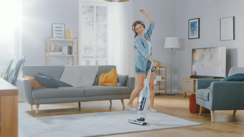 Young Beautiful Woman in Jeans Shirt and Shorts is Listening to Music on Her Headpones, Dancing and Vacuum Cleaning a Carpet in a Cozy Room at Home. She Uses a Cordless Vacuum. She's Happy and Joyful.