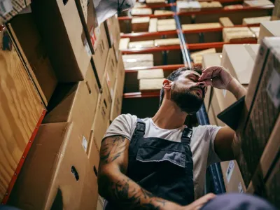 Tattooed bearded worker in overalls having hard day on the work. Storage of import and export firm interior.