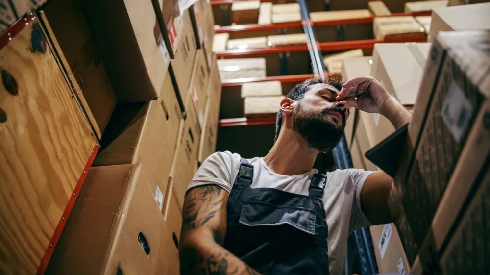 Tattooed bearded worker in overalls having hard day on the work. Storage of import and export firm interior.
