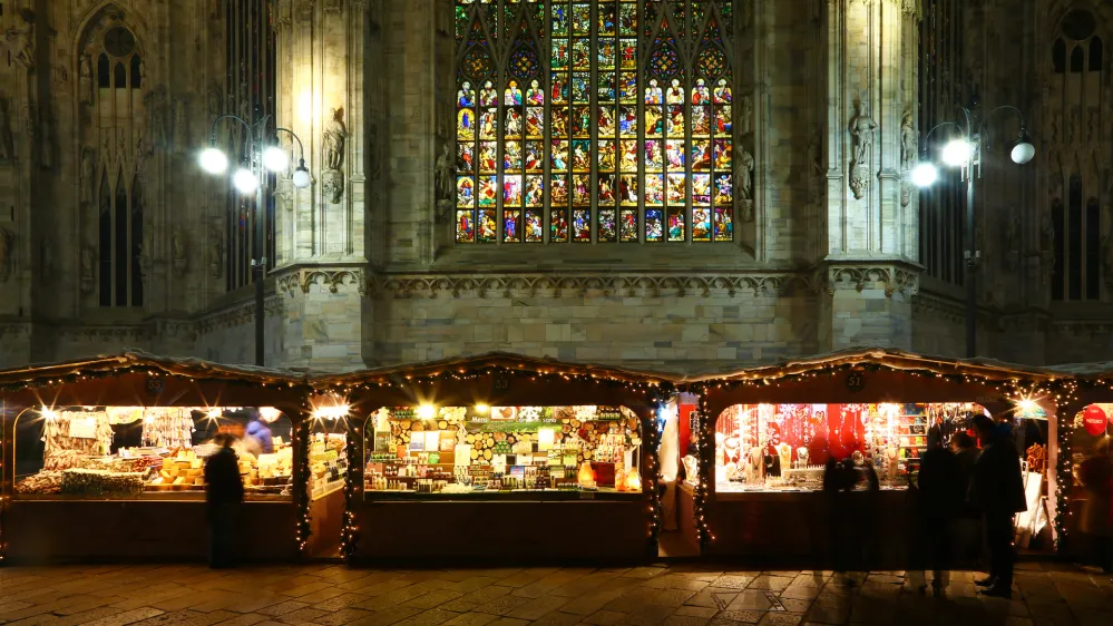 Milan, Italy - December 25, 2015: Christmas market in Piazza del Duomo in late evening. Behind the stalls there is the gothic cathedral with the stained-glass windows lighted for the festivity. Some people are visible in the square.