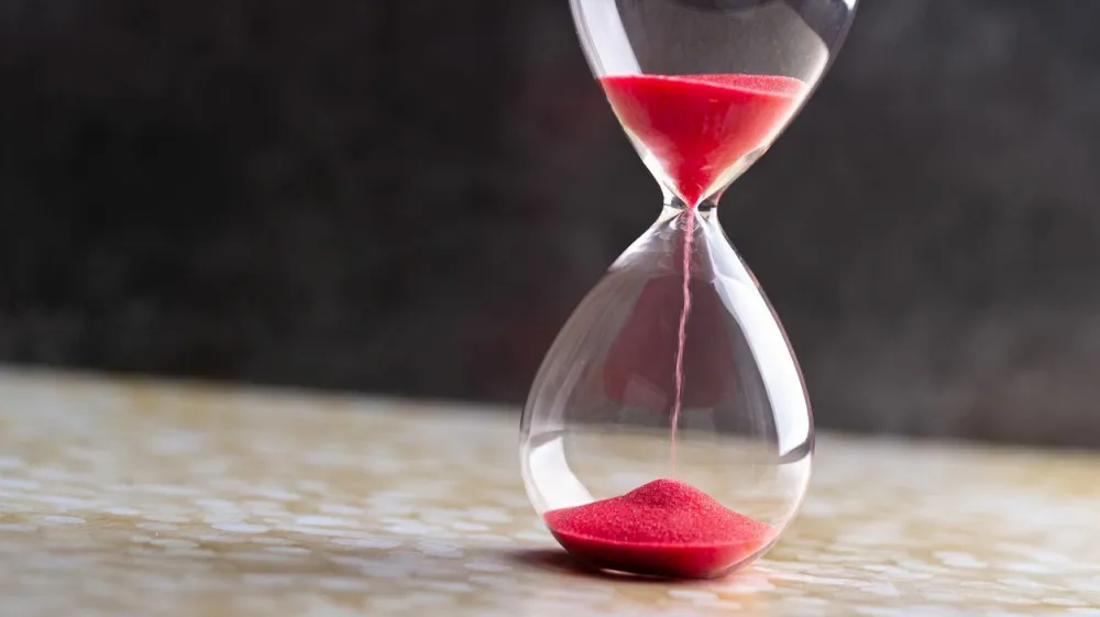 Sand passes through hourglass bulbs measuring the transit time counting down to the deadline, against a dark background