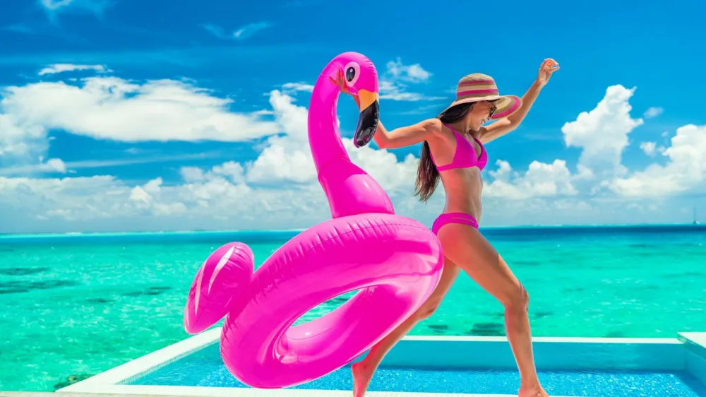 Vacation fun woman in bikini with funny inflatable pink flamingo pool float running of joy jumping by infinity swimming pool. Girl enjoying travel holidays at resort luxury overwater bungalow travel.