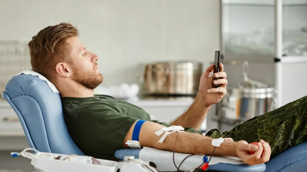 Side view portrait of military man giving blood while laying in chair at blood donation center and watching videos at smartphone