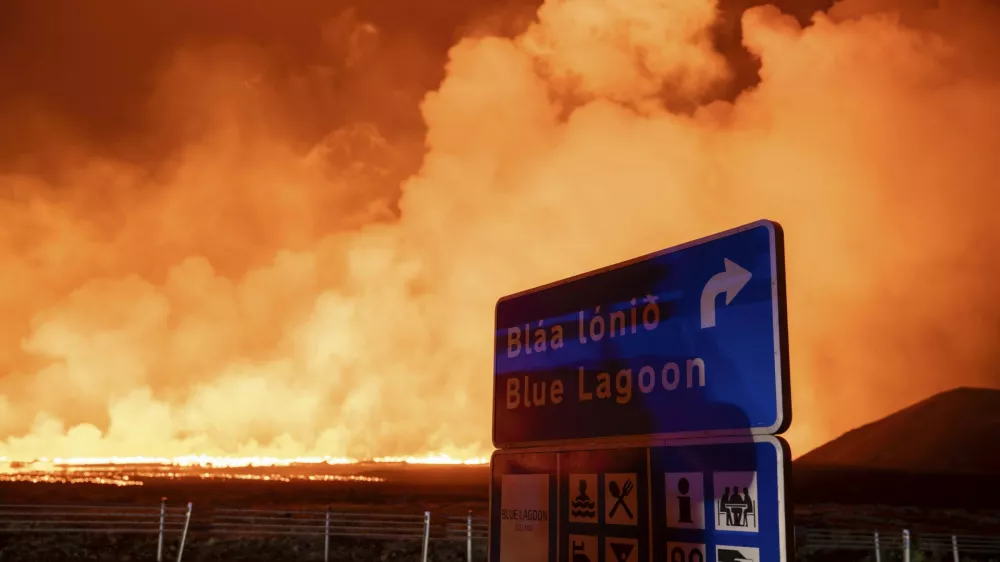 The sign with the direction to the blue lagoon with the new eruption in the background in Grindavik, Iceland, Thursday, Aug. 22, 2024, (AP Photo/Marco di Marco)