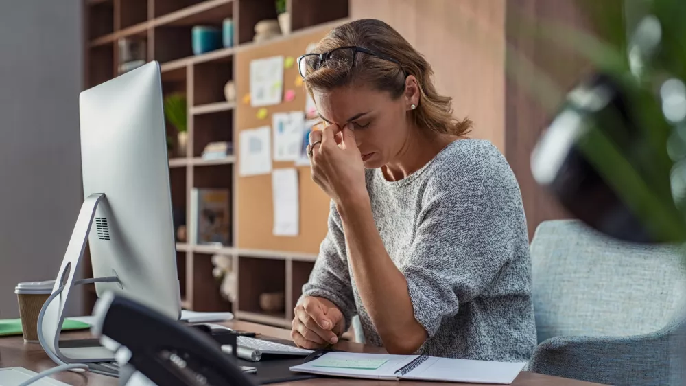 Exhausted businesswoman having a headache in modern office. Mature creative woman working at office desk with spectacles on head feeling tired. Stressed casual business woman feeling eye pain while overworking on desktop computer.