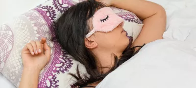 A shallow focus closeup of a female wearing a sleeping eye cover while lying on the bed / Foto: Wirestock