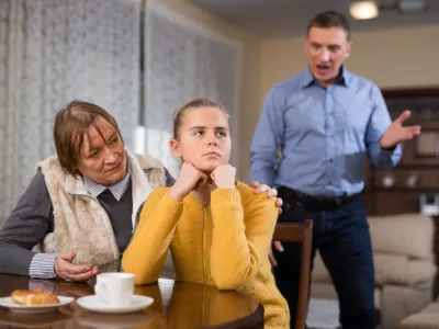Portrait of troubled teen girl sitting at home table, scolded by her father and grandma / Foto: Jackf