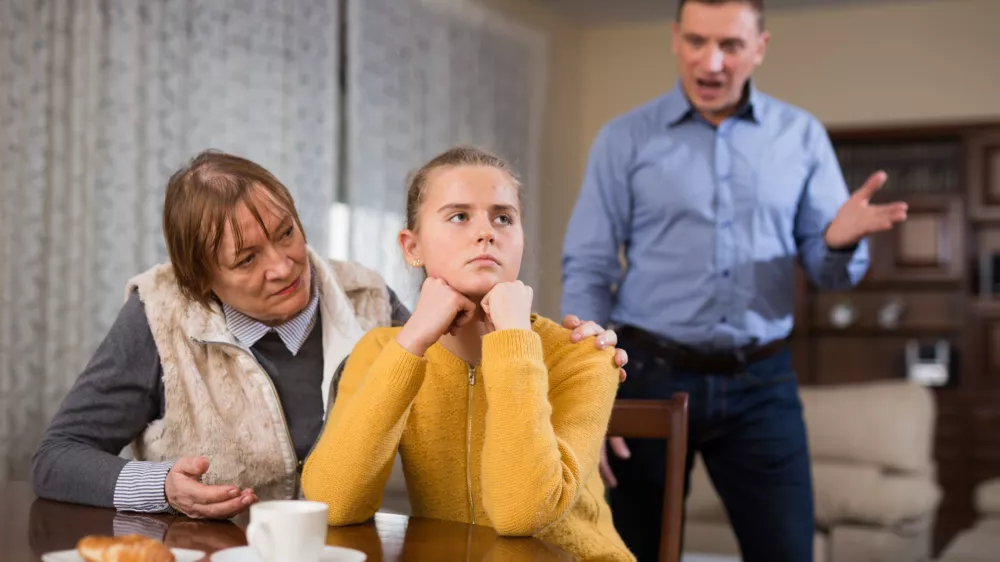 Portrait of troubled teen girl sitting at home table, scolded by her father and grandma / Foto: Jackf