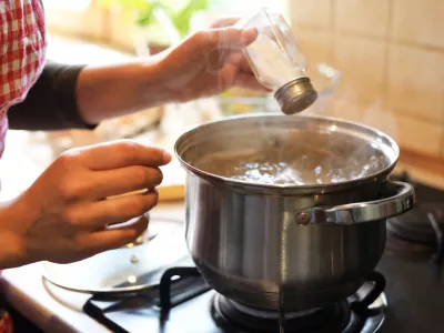 Woman adding salt to boiling water in pot on stove indoors, closeup