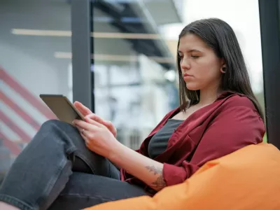 A woman sitting on a beang bag. Her eyes fixed on the screen of her tablet computer. The naturally-lit room surrounds her, with a bookshelf and window visible in the background, creating a sense of comfort and tranquility. / Foto: Bor Kankaras