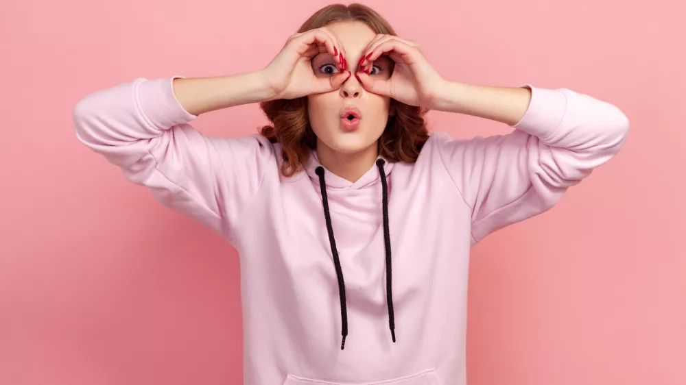 Portrait of amazed teen girl with curly hair in hoodie looking through binoculars gesture and expressing surprise, zooming vision, exploring distance. Indoor studio shot isolated on pink background / Foto: Khosrork