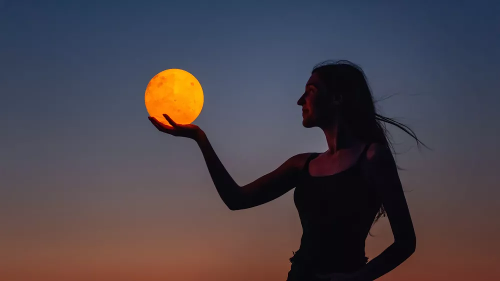 A young woman holds the full moon in her hands against the backdrop of a red sunset. Millennial blogger leads social media. Girl silhouette portrait. Astrology, Waxing Crescent. Copy space / Foto: Julia Pavaliuk