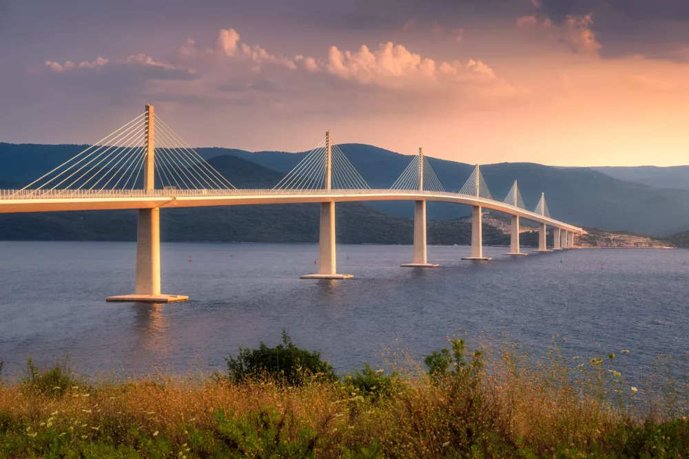 Modern Peljesac bridge and blue sea at colorful sunset in summer in Croatia. Beautiful Peljeski bridge during golden hour, mountains, reflection in water and orange sky with clouds. Architecture