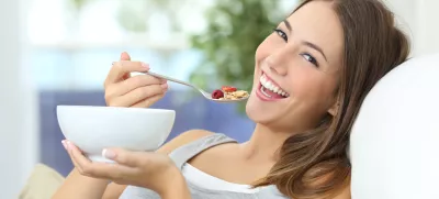 ﻿Happy girl eating cereals with fruits from a bowl sitting on a couch at home