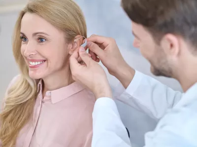 Fitting. Man in a white coat putting hearing aid on ear of smiling middle-aged woman with long blond hair sitting in medical office / Foto: Zinkevych