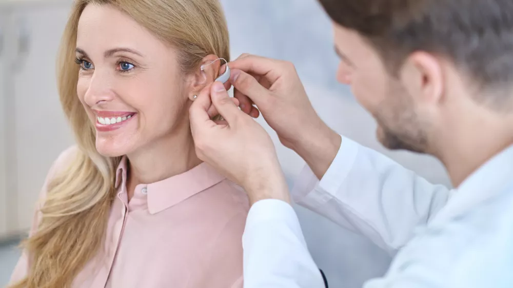 Fitting. Man in a white coat putting hearing aid on ear of smiling middle-aged woman with long blond hair sitting in medical office / Foto: Zinkevych