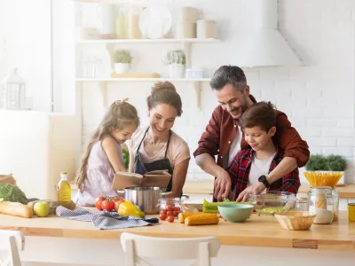 Happy family cooking together on kitchen. Mother and daughter reading recipe to father and son. Dad and boy chopping green vegetable leaf for salad. Home recreation and food preparation on weekend