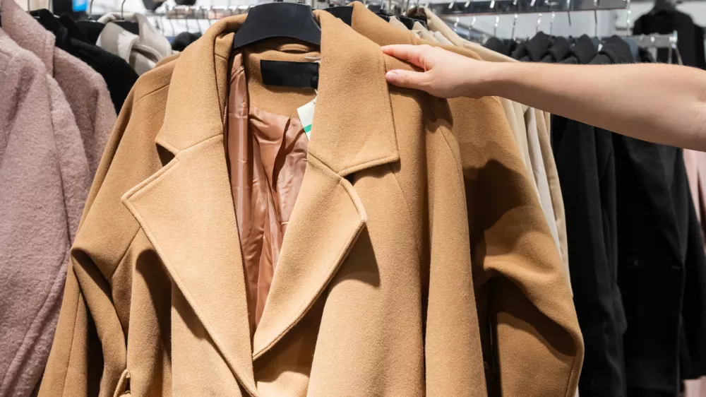 A shopper's hand selecting a stylish camel coat from a rack filled with various garments in a retail clothing store / Foto: Volha Van Meer