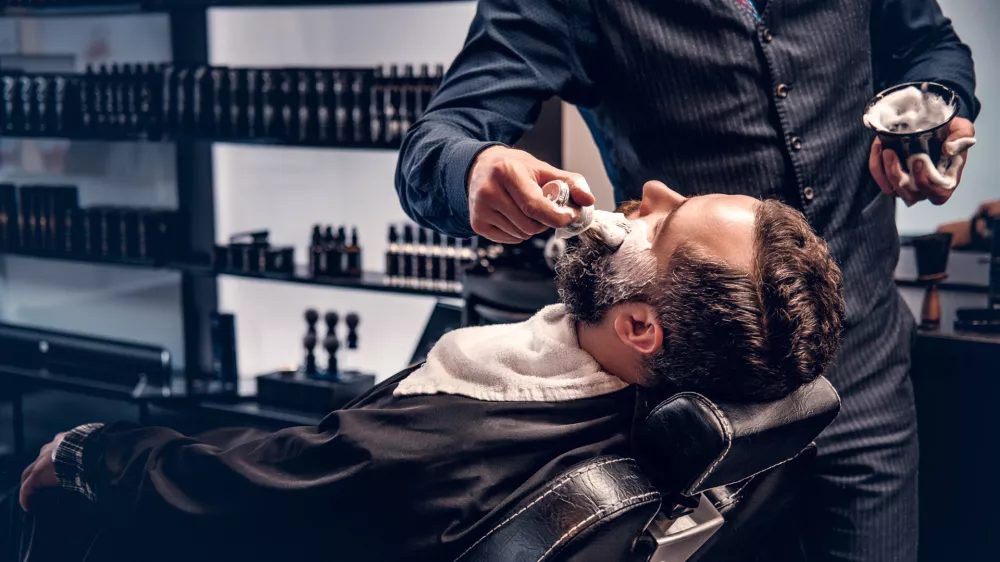 Barber applies shaving foam to a man's face in a saloon. / Foto: Fxquadro