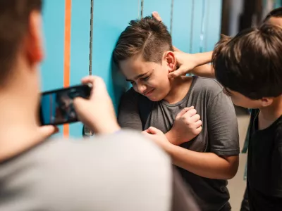 A young boy is being bullied by peers at school. The boy is upset as another boy grabs his neck. A third boy records the bullying incident on his phone. Bullying of teenager in high scool.