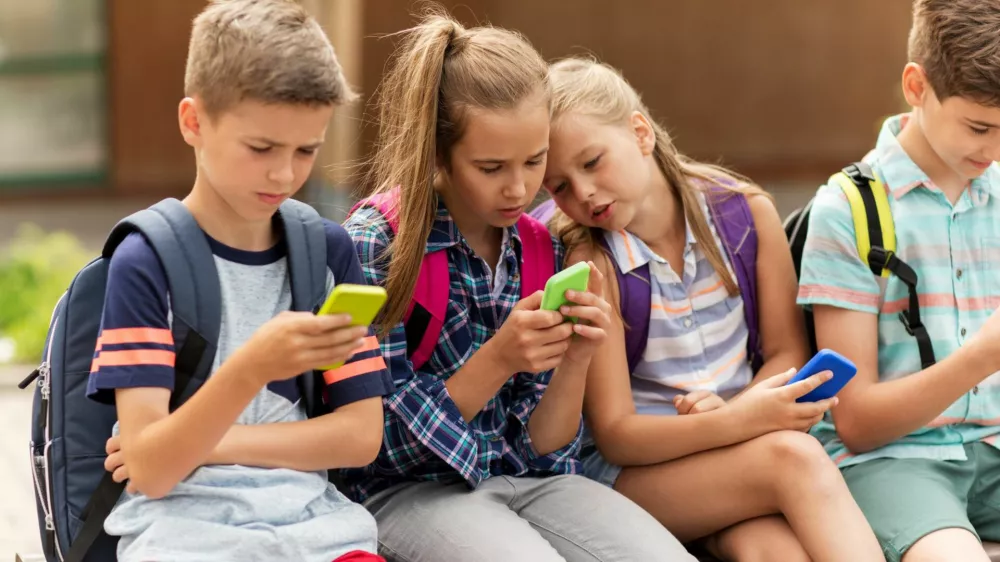 ﻿primary education, friendship, childhood, technology and people concept - group of happy elementary school students with smartphones and backpacks sitting on bench outdoors