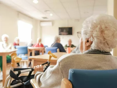 Back view of senior woman with short white hair sitting on chair with foldable walker with friends in blurred background at nursing home / Foto: Daniel Megias