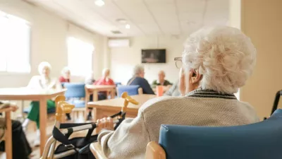 Back view of senior woman with short white hair sitting on chair with foldable walker with friends in blurred background at nursing home / Foto: Daniel Megias