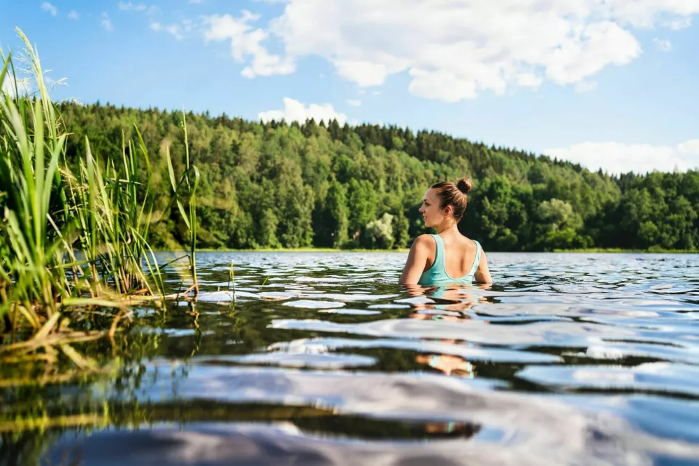 Swimming in lake in Finland. Woman in summer at beach. Finnish bathing in water in nature. Back view of person. Blue sky and beautiful green forest. Relax in Scandinavia after cottage sauna. / Foto: Tero Vesalainen