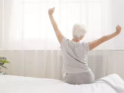 Senior woman sitting on her bed in morning, stretching with arms raised, back view, free space / Foto: Prostock-studio
