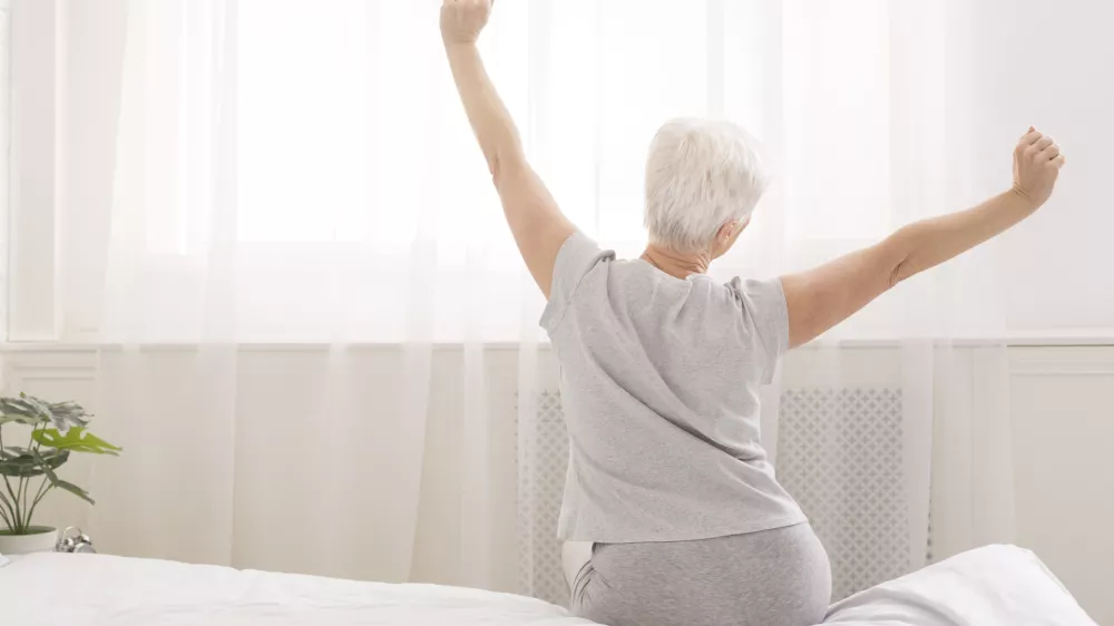 Senior woman sitting on her bed in morning, stretching with arms raised, back view, free space / Foto: Prostock-studio
