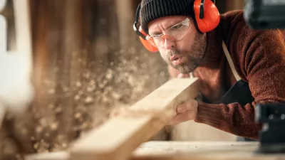Adult male woodworker in protective goggles and headphones blowing sawdust from wooden detail while working in carpentry workshop / Foto: Evgenyatamanenko