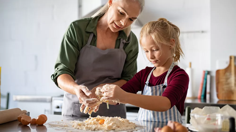 Cute girl learning preparing dough with mature grandmother in kitchen. Lovely old grandma and granddaughter preparing cookies with flour and eggs on kitchen counter. Senior granny in apron teaching her child to make cookies or pie. / Foto: Ridofranz