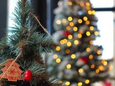 A decorated Christmas tree branch featuring a small wooden tree ornament and bright red berry detail, set against a softly blurred background of warm glowing lights. The bokeh highlights classic seasonal charm, handcrafted holiday style, and cozy winter ambiance. Perfect for festive campaigns, December storytelling, interior Christmas promotion, greeting card visuals, and winter celebration mood boards