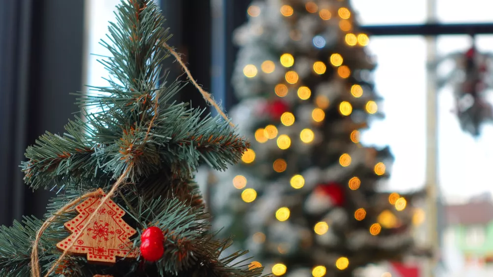 A decorated Christmas tree branch featuring a small wooden tree ornament and bright red berry detail, set against a softly blurred background of warm glowing lights. The bokeh highlights classic seasonal charm, handcrafted holiday style, and cozy winter ambiance. Perfect for festive campaigns, December storytelling, interior Christmas promotion, greeting card visuals, and winter celebration mood boards