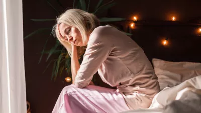 Upset middle-aged woman sit on bed in bedroom lost in thoughts thinking pondering of problem solution, depressed unhappy mature female look in distance suffer from depression, having personal problems