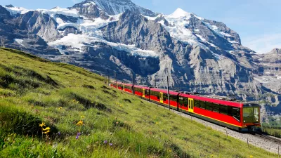 A tourist train travels on Jungfrau Railway from Jungfraujoch (Top of Europe) to Kleine Scheidegg & wild flowers bloom on a green grassy hillside under blue sunny sky in Bernese Oberland, Switzerland