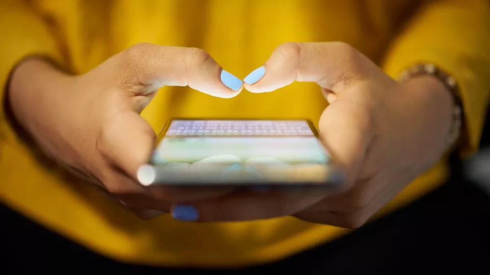 Young woman using cell phone to send text message on social network at night. Closeup of hands with computer laptop in background