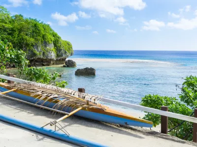 Out-rigger canoe under palm frond to shield some rainwater from boat on ledge overlooking scenic coastline and vast Pacific Island, Niue.
