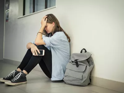 Upset and depressed girl holding smartphone sitting on college campus floor holding head. University sad student suffering from depression sitting on floor at high school. Lonely bullied teen in difficulty with copy space. / Foto: Ridofranz