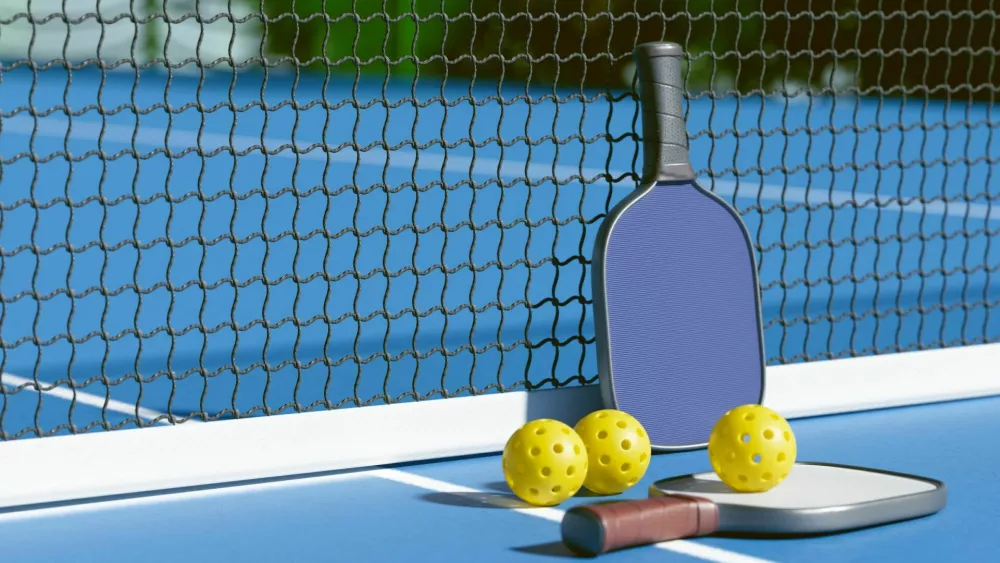 A pickleball paddle and balls resting by the net on a blue court, ready for a game. 3d rendering / Foto: Olga Nikiforova