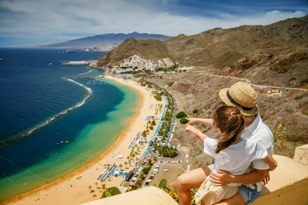 Mother and daughter sitting on a ledge, looking out over Las Teresitas beach in Tenerife, pointing towards the beach. Traveling with children concept. / Foto: Anita_bonita