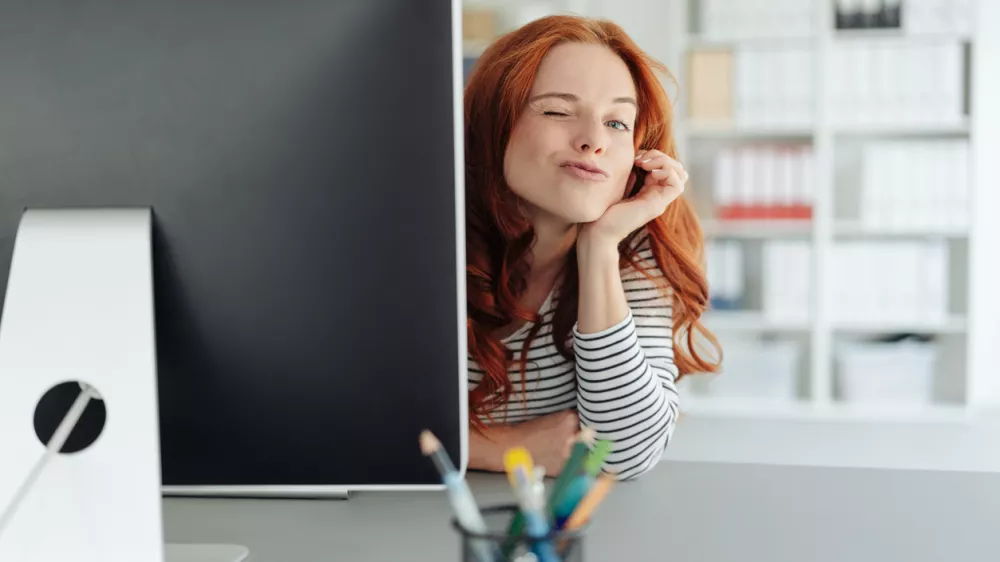 Playful young businesswoman winking at the camera as she peers around the edge of a large desktop monitor in a spacious office