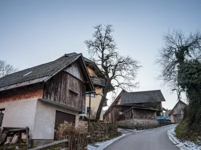 In the heart of Stara Fuzina, wooden and masonry chalets cluster along a winding lane. Textured facades, steep gabled roofs, and small windows reveal the practical alpine architecture developed for cold seasons and mountain life. Weathered boards and stone retaining walls add a sense of age and continuity, while bare trees and leftover snow signal winter. The composition highlights everyday rural character rather than tourist icons, making it ideal for illustrating Slovenian village life, vernacular building styles, heritage preservation, and the human scale of communities near Lake Bohinj. / Foto: Balkanscat Getty Images