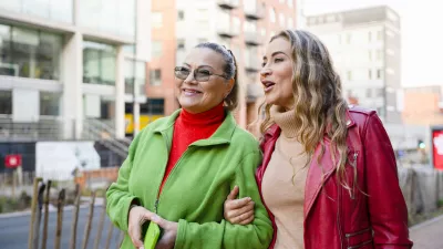 Two women walk side by side on a city street. They are smiling and talking, enjoying the sunny day among buildings and shops. The street is lively and active. / Foto: Iryna Melnyk Getty Images