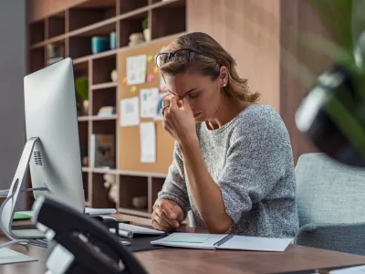Exhausted businesswoman having a headache in modern office. Mature creative woman working at office desk with spectacles on head feeling tired. Stressed casual business woman feeling eye pain while overworking on desktop computer.