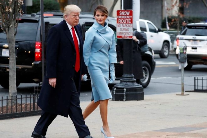 Donald Trump je oblečen v plašč in obleko temne barve z rdečo kravato, medtem ko je Melania v kostimu nebeško modre barve....