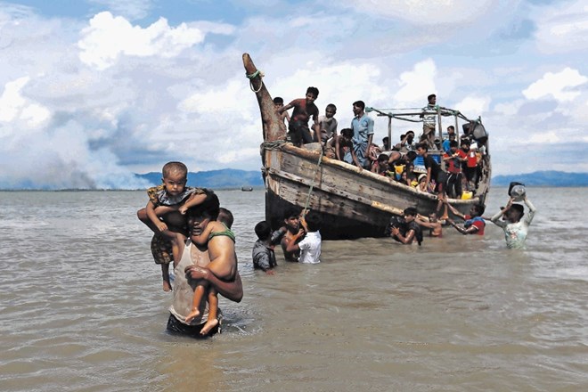 Begunci iz ljudstva  Rohinga v Bengalskem zalivu z dimom v ozadju, kjer gorijo njihove domačije v Mjanmaru.
